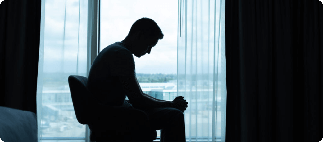 Silhouette of a person sitting alone by a window, head bowed in a posture suggesting depression or contemplation