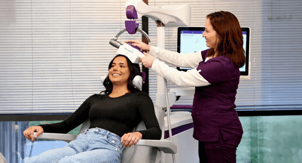 A woman sitting calmly in a treatment chair while a medical technician positions a NeuroStar TMS coil against the left side of her head.