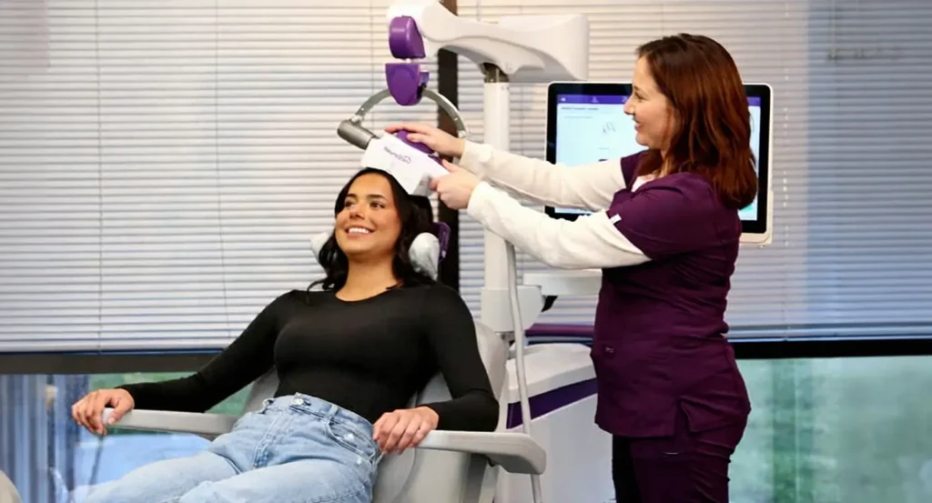 A woman sitting calmly in a treatment chair while a medical technician positions a NeuroStar TMS coil against the left side of her head.