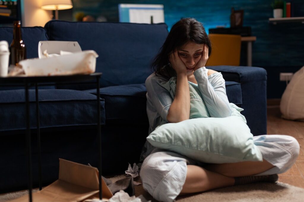 A distressed woman sitting cross-legged on the floor of a dark, cluttered living room, clutching a pillow and holding her face in her hands.