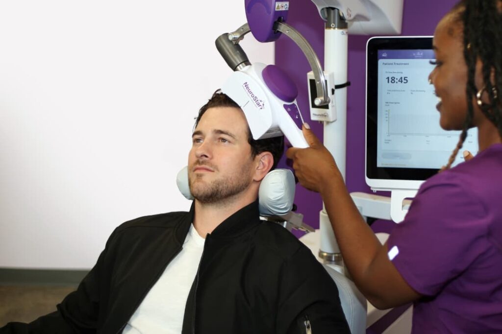 Clinician operating a NeuroStar device on a seated patient during a TMS therapy for depression vs medication session.